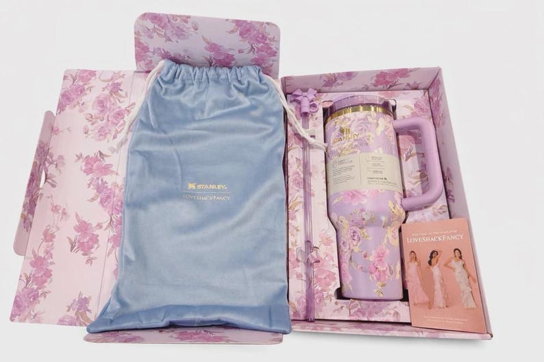 May include: A lavender Stanley tumbler with a floral pattern, a matching straw, and a light blue drawstring pouch inside a floral gift box. The box includes a card featuring three people in dresses and the text LoveShackFancy and Stanley.