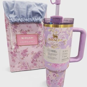 May include: A lavender Stanley and LoveShackFancy tumbler with a floral pattern, a matching handle, and a straw topped with a bow accessory. Beside it sits a coordinating floral gift bag with a light blue fabric top and a Stanley LoveShackFancy label.