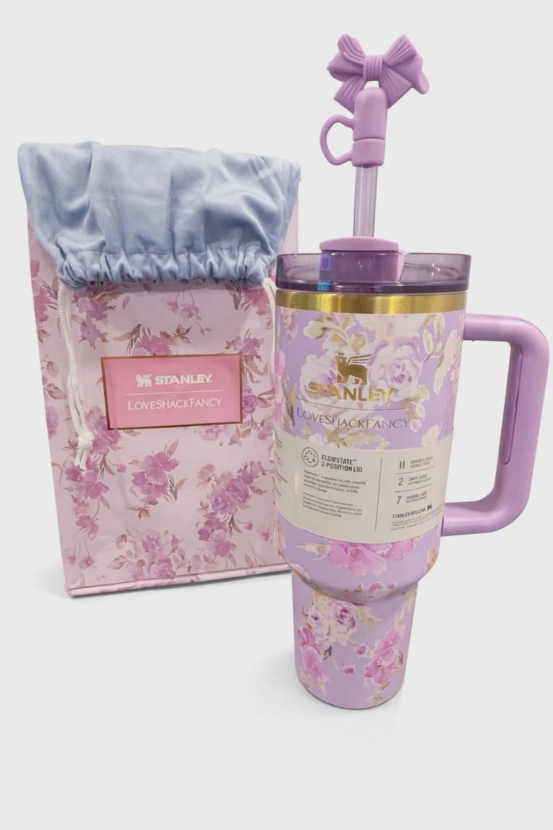 May include: A lavender Stanley and LoveShackFancy tumbler with a floral pattern, a matching handle, and a straw topped with a bow accessory. Beside it sits a coordinating floral gift bag with a light blue fabric top and a Stanley LoveShackFancy label.