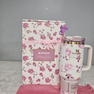 May include: A white Stanley Quencher tumbler and matching box featuring a pink rose floral pattern. The tumbler has a handle, a clear lid, and a label reading Stanley LoveShackFancy. A small purple bow accessory is attached to the box.