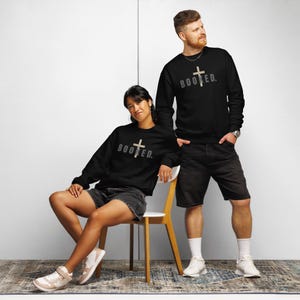 Op de afbeelding: Zwarte crewneck sweatshirts met het woord "BOOKED" en een kruisafbeelding in een lichte kleur. De sweatshirts worden gedragen met zwarte shorts en witte sneakers. De afbeelding toont twee personen, één zittend en één staand.