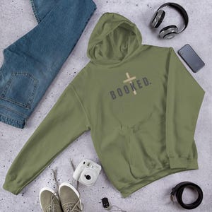 Op de afbeelding: Een olijfgroene hoodie met het woord "BOOKED" op de voorkant, met een kruisontwerp. De hoodie ligt met een blauwe jeans, een koptelefoon, een smartphone, een camera, schoenen en een riem.
