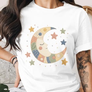 May include: White t-shirt featuring a whimsical design of a crescent moon with a smiling face, surrounded by colorful stars. The moon is composed of various patchwork patterns in pastel colors. A charming and dreamy graphic tee.