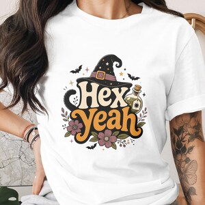Könnte beinhalten: Weißes T-Shirt mit einem farbenfrohen Halloween-Motiv. Das Design zeigt einen Hexenhut, Fledermäuse, Blumen und die Worte "Hex Yeah" in einer Retro-Schriftart. Eine Zaubertrankflasche ist ebenfalls enthalten.