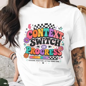 Op de afbeelding: Wit T-shirt met een kleurrijke afbeelding met de tekst "CONTEXT SWITCH IN PROGRESS". Het ontwerp bevat bloemen, een bliksemschicht en een voortgangsbalk. De tekst "Focusing. Resetting. Getting things done." is ook opgenomen.