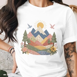 May include: White t-shirt with a colourful mountain landscape design. The graphic includes a yellow sun, blue and pink birds, and floral and tree elements. The mountains are composed of patchwork patterns.