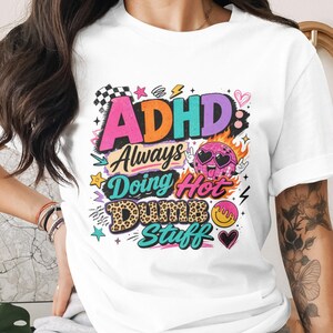 May include: White t-shirt with a colorful graphic design. The design includes the text "ADHD Always Doing Hot Dumb Stuff" in various fonts and colors, along with cartoon illustrations of a disco ball, lightning bolts, and hearts.