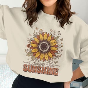 以下が含まれることがあります： クリーム色のスウェットシャツ。大きなヒマワリのデザインと「Chase the Sunshine」の文字入り。ヒマワリは黄色、茶色、オレンジ色で、蝶と花のアクセントが施されています。カジュアルで快適なトップス。