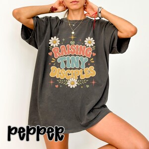 May include: A charcoal gray short-sleeve t-shirt featuring a graphic print with the text "RAISING TINY DISCIPLES" in a retro font. The design includes white daisies, small red hearts, and star accents in a muted color palette of pink, teal, and yellow.