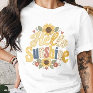 May include: White t-shirt featuring the words "Hello Sunshine" in a colorful, patterned font, surrounded by sunflowers and floral accents. The design includes yellow, blue, pink, and green elements, creating a cheerful and summery vibe.