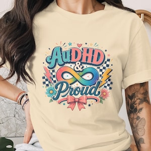 Puede incluir: Camiseta color crema con un gráfico colorido que dice "AuDHD & Proud". El diseño incluye un símbolo de infinito, rayos, flores y un lazo. La camiseta es de estilo casual.