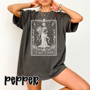 May include: A charcoal grey short-sleeve t-shirt featuring a white graphic print of a skeleton dressed in a gown holding a bouquet and a book. The design is framed like a tarot card with floral borders, stars, and the text THE MAID OF HONOR.