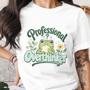 May include: White t-shirt with a cartoon frog and the words "Professional Overthinker." The design incorporates green leaves, a yellow flower, and a pink heart. The frog is light green and looks thoughtful.