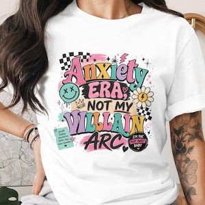 Può includere: T-shirt bianca con un design grafico colorato. Il design presenta il testo "Anxiety Era, Not My Villain Arc" in vari font e colori, insieme a una faccina sorridente, una margherita e altri elementi decorativi. La maglietta ha maniche corte.