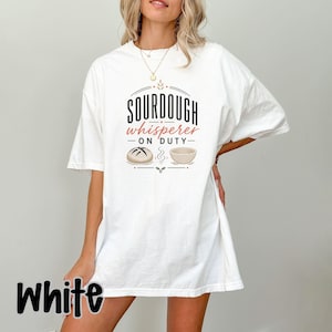Könnte beinhalten: Ein weißes Kurzarm-T-Shirt mit dem schwarzen und terrakottafarbenen Schriftzug SOURDOUGH whisperer ON DUTY. Die Grafik enthält eine Strichzeichnung eines runden Brotlaibs und eines Gärkörbchens, mittig auf der Brust des Kleidungsstücks.