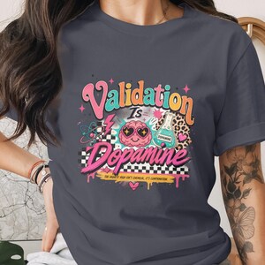 Può includere: T-shirt grigio scuro con un design grafico colorato. Il design presenta le parole "Validation is Dopamine" in un font retrò, insieme a un cervello con occhi a forma di cuore, un becher scientifico e altri elementi. La maglietta ha maniche corte.