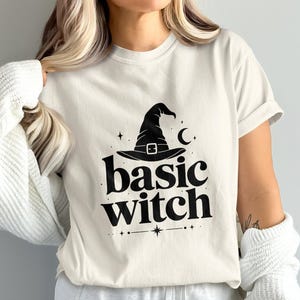 May include: A cream-colored short-sleeve t-shirt featuring a black graphic print on the chest. The design includes a witch hat, a crescent moon, small stars, and the text "basic witch" in a bold, stylized serif font centered below the hat.