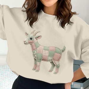 May include: Cream-colored sweatshirt featuring a patchwork goat design in pink and green. The goat has horns and is made of various fabric patterns. The sweatshirt has long sleeves and a crew neck.