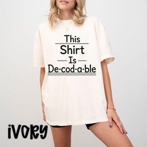 Op de afbeelding: Een ivoorkleurig t-shirt met korte mouwen en zwarte tekst die This Shirt Is De-cod-a-ble leest. Het ontwerp bevat horizontale lijnen boven en onder de tekst, waarbij het woord decodable in lettergrepen is verdeeld met punten ertussen.