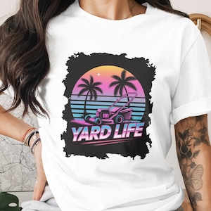 Puede incluir: Camiseta blanca con un diseño retro que presenta una cortadora de césped, palmeras y una puesta de sol. Las palabras "YARD LIFE" se muestran en negrita, ideal para los amantes del jardín.