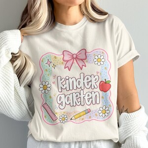 May include: A cream-colored t-shirt featuring a graphic with the text kindergarten in a bubble font. The design includes a pink bow, a red apple, a yellow pencil, a pink ruler, and white daisies, all enclosed within a scalloped pastel border.