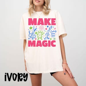 May include: An ivory short-sleeve t-shirt featuring the pink text "MAKE MAGIC" and a central graphic of a blue pair of scissors, a green spool of thread, and a blue paintbrush with the text "Creativity lives here" in blue script.