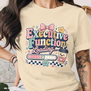May include: A cream-coloured t-shirt featuring the phrase "Executive Function Loading" in a retro font. The design includes a pink bow, stars, a coffee cup, and a loading bar. A fun, whimsical graphic tee.