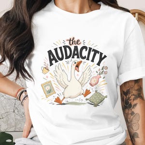 May include: White t-shirt featuring a cartoon goose with the text "the AUDACITY". The goose is running with its mouth open, surrounded by decorative elements like a teacup, flowers, and a framed picture. The design has a whimsical, illustrative style.