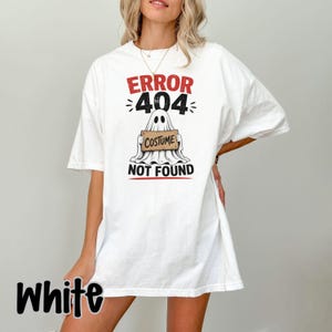 May include: A white short-sleeve t-shirt featuring a graphic of a ghost holding a sign that reads COSTUME. Above the ghost, the text reads ERROR 404, and below the ghost, the text reads NOT FOUND. The design uses red and black lettering.