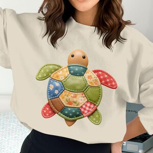 May include: Cream-colored sweatshirt featuring a colorful patchwork turtle design. The turtle has a wooden head and multi-colored shell and flippers. The design includes floral, polka dot, and gingham patterns.