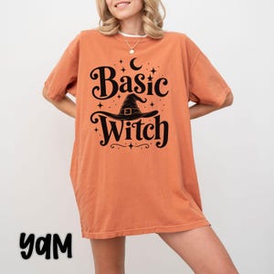 Può includere: Una t-shirt a maniche corte color arancione bruciato con una stampa grafica nera. Il design include il testo "Basic Witch" in un carattere stilizzato, centrato sopra e sotto l'illustrazione di un cappello da strega, circondato da stelle e una luna.