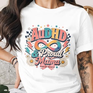 Puede incluir: Camiseta blanca con la frase "AuDHD & Proud Mama" en un diseño retro colorido. El gráfico incluye un símbolo de infinito, rayos, margaritas, estrellas y un lazo rosa. El texto está en una fuente colorida.