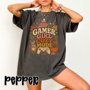 Op de afbeelding: Een antracietgrijs t-shirt met korte mouwen en een grafische print van een gebreide muts, een dampende mok, een gamecontroller en een pompoen. De tekst "GAMER GIRL COZY MODE" staat in een retro lettertype op het shirt.