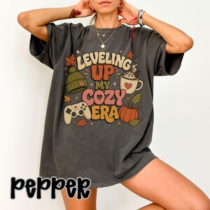 Op de afbeelding: Een antracietgrijs T-shirt met korte mouwen en een grafische print met de tekst "LEVELING UP MY COZY ERA". Het ontwerp bevat een groene gebreide muts, een mok warme chocolademelk met een hartje, een gamecontroller en een oranje pompoen.