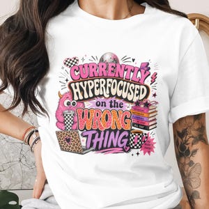 May include: White t-shirt with a colorful graphic that reads "CURRENTLY HYPERFOCUSED on the WRONG THING." The design includes a pink monster, books, a laptop, and coffee cups. The shirt is a casual, everyday wear item.