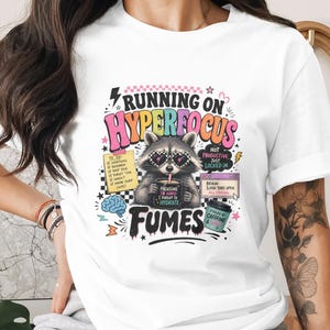 May include: White t-shirt with a colorful graphic featuring a raccoon wearing sunglasses and holding a mug. The text reads "Running on Hyperfocus Fumes" with additional text and illustrations.