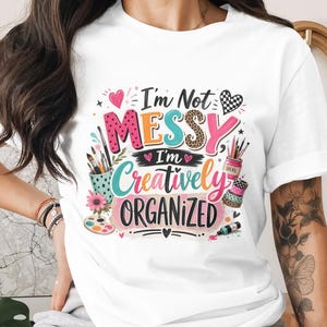 Könnte beinhalten: Weißes T-Shirt mit dem Text "I'm Not Messy I'm Creatively Organized" in einem farbenfrohen Design. Das Design umfasst Pinsel, Farbpaletten und andere Kunstutensilien, was auf ein künstlerisches Thema hindeutet.