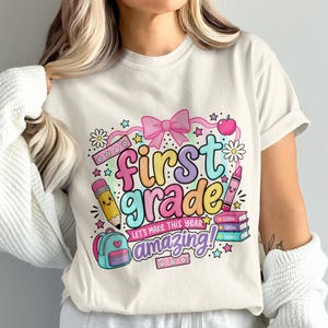May include: A cream-colored t-shirt featuring a colorful graphic with the text "first grade" and "LET'S MAKE THIS YEAR AMAZING!". The design includes a pink bow, a pencil, a crayon, a backpack, an apple, and a stack of books with "LEARN GROW SHINE".