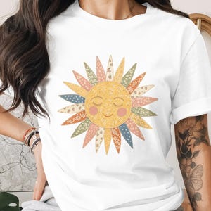 May include: White t-shirt featuring a smiling sun graphic. The sun has a yellow face with closed eyes and rosy cheeks. Its rays are composed of various patterned fabrics in pastel colors, creating a cheerful and whimsical design. The shirt is a casual wear item.