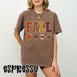 May include: A brown short-sleeve t-shirt featuring the word FALL in large block letters. Below the text are graphic icons of an orange pumpkin, a brown maple leaf, a coffee cup with a heart, and a brown football, all in a distressed style.
