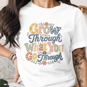 May include: White t-shirt with the text "Grow Through What You Go Through" in a colorful, retro font. The design includes floral accents in yellow, blue, pink, and green. A casual, inspirational tee.