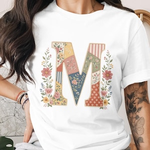 May include: White t-shirt with a large, colourful letter "M" in a patchwork design. The letter is framed by floral and leaf accents. The shirt is a casual, everyday wear item.