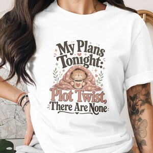 Puede incluir: Camiseta blanca con un gráfico que presenta un gato bajo una manta sosteniendo una taza. El texto dice "My Plans Tonight: Plot Twist, There Are None" en una fuente caprichosa. El diseño incluye corazones y detalles florales.