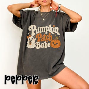 Può includere: Una t-shirt a maniche corte grigio antracite con una stampa grafica che riporta il testo Pumpkin Patch Babe in crema e arancione. Il design include un fantasma dei cartoni animati che tiene una tazza di caffè e una zucca arancione sorridente.