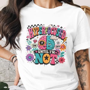 May include: White t-shirt with a colorful graphic design. The design features a split brain illustration, one side pink and the other blue, with the words "HYPERFOCUSED" and "UNTIL I'M NOT". Decorative elements include flowers, hearts, and a lightning bolt.