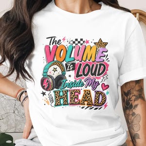 Puede incluir: Camiseta blanca con un gráfico colorido que dice "The Volume Is Loud Inside My HEAD". El diseño incluye auriculares, estrellas y un estampado de leopardo. Una prenda de vestir casual.