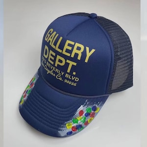 May include: Navy blue trucker hat with the text "GALLERY DEPT." in gold lettering. The brim features colorful paint splatters. The hat includes the address 7613 Beverly Blvd, Los Angeles, Ca. 90036.