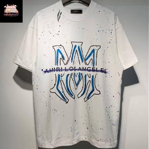 May include: White t-shirt with a blue and black design. The shirt features a large, stylized "M" logo with "AMIRI LOS ANGELES" printed below. The shirt has a splatter paint effect.