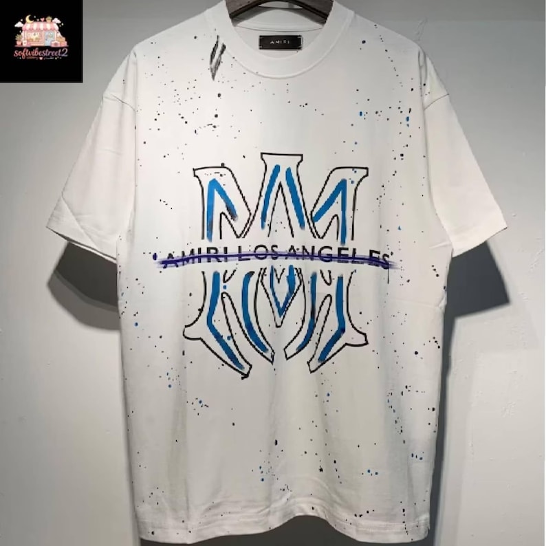 May include: White t-shirt with a blue and black design. The shirt features a large, stylized "M" logo with "AMIRI LOS ANGELES" printed below. The shirt has a splatter paint effect.