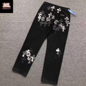 Op de afbeelding: Zwarte jeans met kruis- en logo-details. De jeans met rechte pijpen hebben zilveren en zwarte kruisapplicaties. Er zit een label aan de achterkant. De broek is ongeveer 100 cm lang.